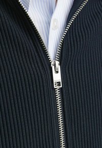 Marineblaue gerippte Zip-Jacke mit silbernem Reißverschluss und einem weißen Hemd darunter, das blaue vertikale Streifen zeigt.