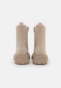 Beige ruskindsankelstøvler med en tekstureret gummisål, lynlås lukning og en pull-tab bagpå; enkelt design med en kraftig silhuet.