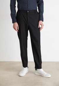 Svarte bukser med en skreddersydd passform, med elastisk linning og knappelukking, kombinert med hvite joggesko og en mønstrete skjorte.