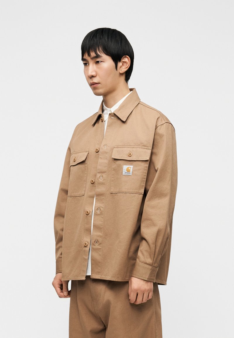 Brown cotton overshirt with a button front, two chest pockets, and a logo patch. The design features long sleeves and a relaxed fit.