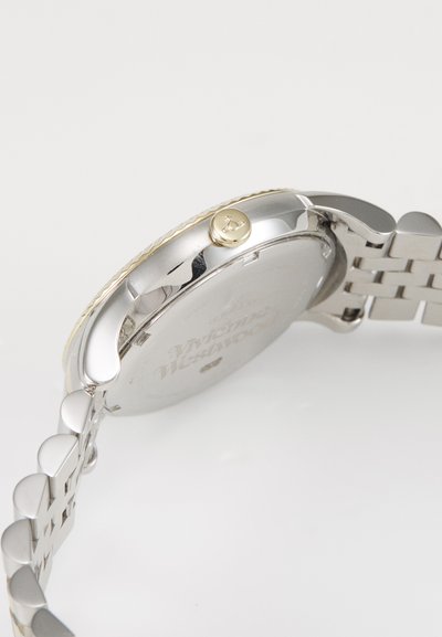 Hõbedane metallkell ümara korpusega, kuldsete rõngastega kroon ja kootud käevõru, millel on poleeritud lingid ja sile tekstuur.