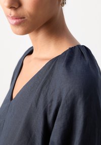 Gros plan d'une femme portant un haut bleu marine à col en V et une boucle d'oreille créole dorée avec un détail texturé sur fond blanc.