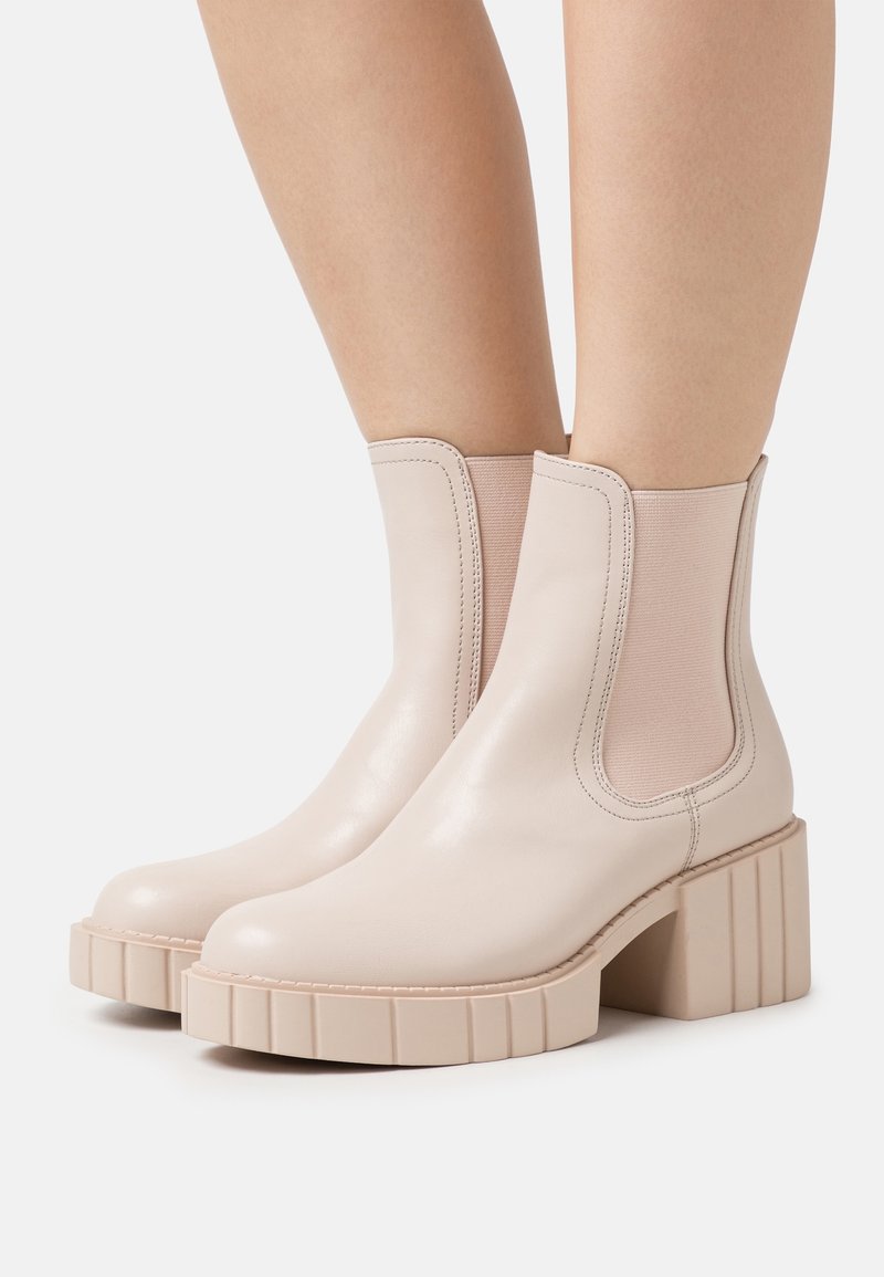 Bottines beiges avec semelles épaisses et panneaux latéraux élastiques portées sur des jambes nues sur un fond uni.