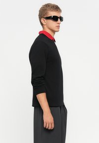 Young man in black long-sleeve shirt with red collar, dark gray pants, and black sunglasses, standing sideways against a white background.