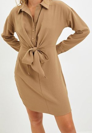 Femme portant une robe-chemise marron clair à manches longues, nouée devant, debout les mains sur les hanches devant un fond uni.