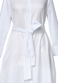 Valkoinen shirt dress, joka on valmistettu tekstuurista kankaasta. Tässä on napitettava etuosa, pitkät hihat ja sidottu satiinivyö, joka korostaa vyötäröä.