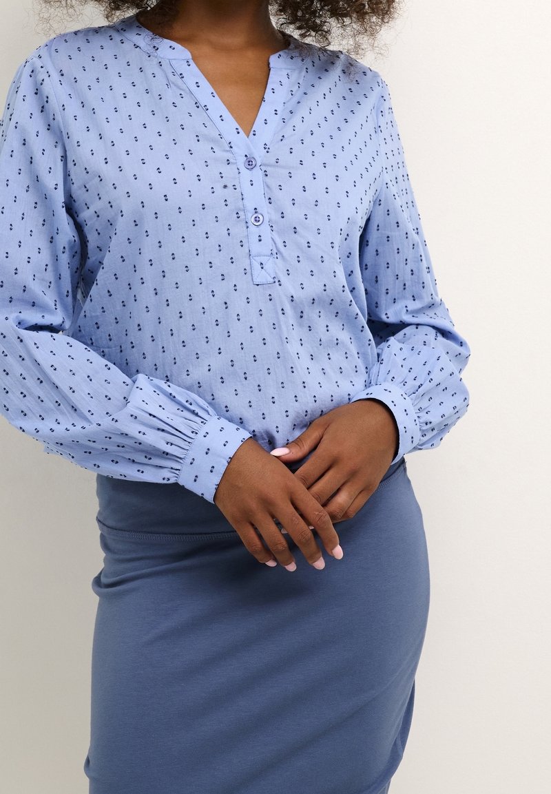 Blouse bleu clair avec petits motifs bleu foncé, une patte de boutonnage et des manches froncées, associée à une jupe bleu ajustée. Texture de tissu douce.