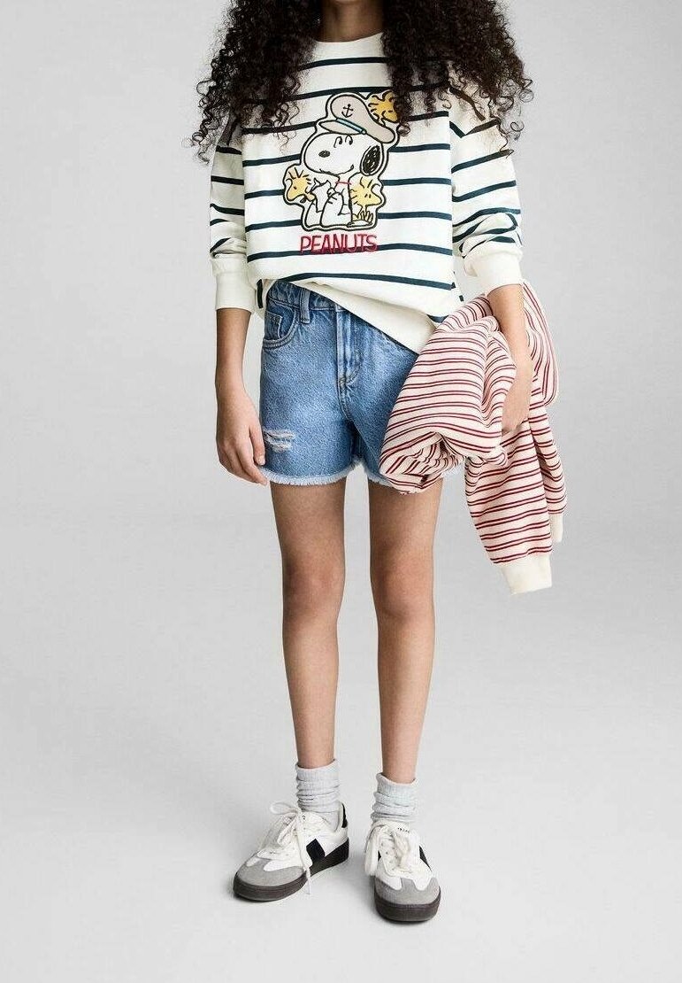 Enfant portant un sweat-shirt Peanuts rayé, un short en jean, des baskets blanches, tenant un pull rayé rouge et blanc dans une main.