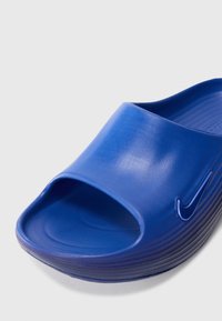 Sandale de sport bleue avec un design ouvert à l'avant, texture lisse et brillante, et semelle crantée. Présente un petit logo en accent sur le côté.
