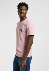 T-shirt in cotone rosa chiaro con maniche corte, colletto a giro e logo nero "Lee" sul petto. Indossato con jeans blu, che mostrano una vestibilità rilassata.