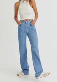 Jean bleu clair taille haute avec jambes larges, poches avant classiques et fermeture à glissière. Assorti à un haut blanc sans manches moulant et des baskets.