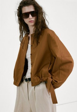 Mujer con cabello largo y rizado, con grandes gafas de sol oscuras, chaqueta marrón abotonada, blusa blanca, pantalones beige y cinturón negro con hebilla dorada.