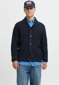 Donkerblauwe gebreide cardigan met een sjalkraag, voorzien van textuurpatronen, vijf knopen en geribbelde manchetten, gedragen over een spijkerhemd.