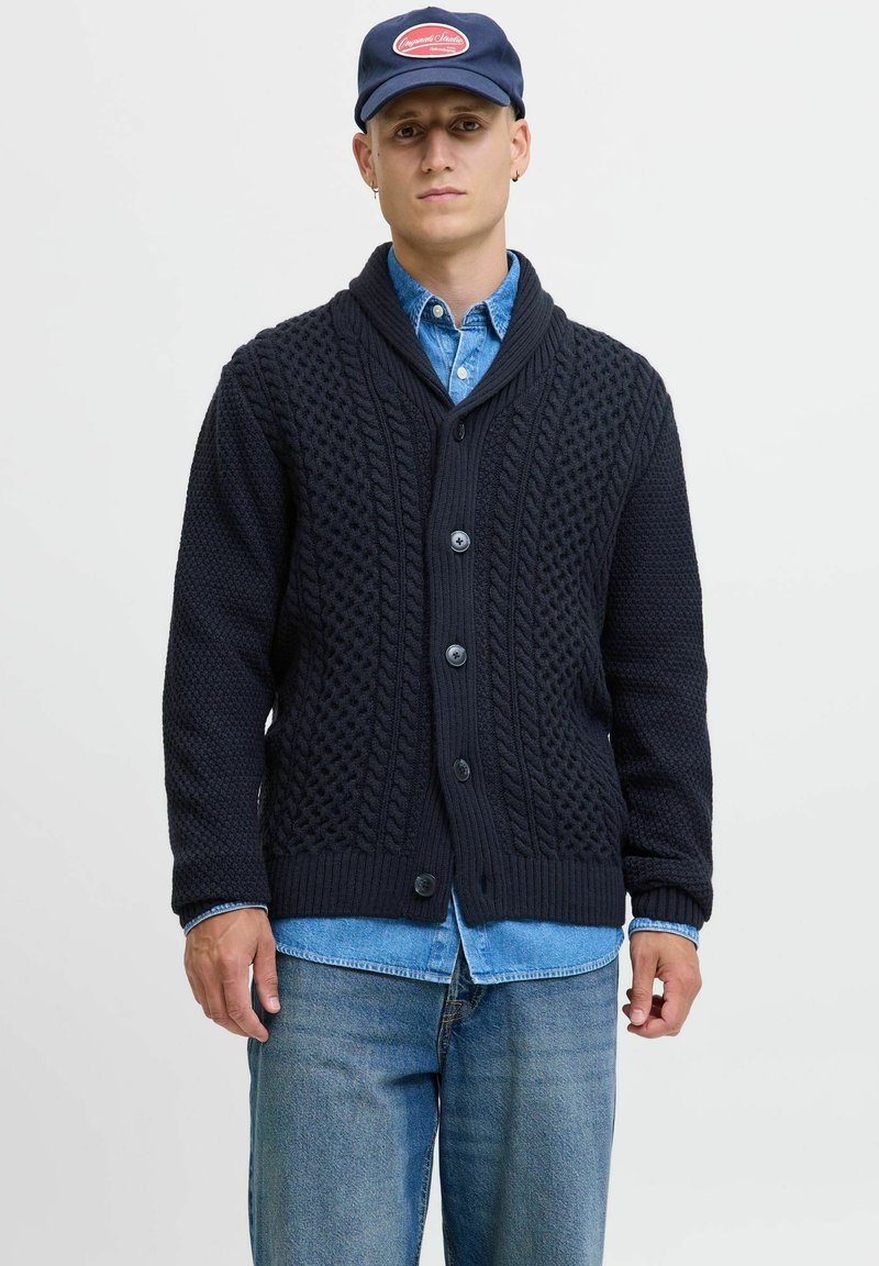 Donkerblauwe gebreide cardigan met een sjalkraag, voorzien van textuurpatronen, vijf knopen en geribbelde manchetten, gedragen over een spijkerhemd.