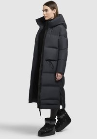 Abrigo largo acolchado en gris oscuro con capucha, que presenta un cierre delantero, cintura entallada y bolsillos laterales; combinado con zapatillas negras de caña alta.
