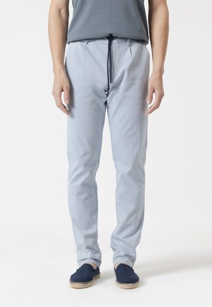 Hombre con pantalones claros con cordón y zapatos tipo slip-on azul oscuro, de pie frente a un fondo blanco.