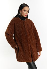 Brun, fluffy, tekstureret frakke med løs pasform, rund halsudskæring og sidelommer. Bæres over en sort turtleneck. Glat, skinnende stof i bunden.