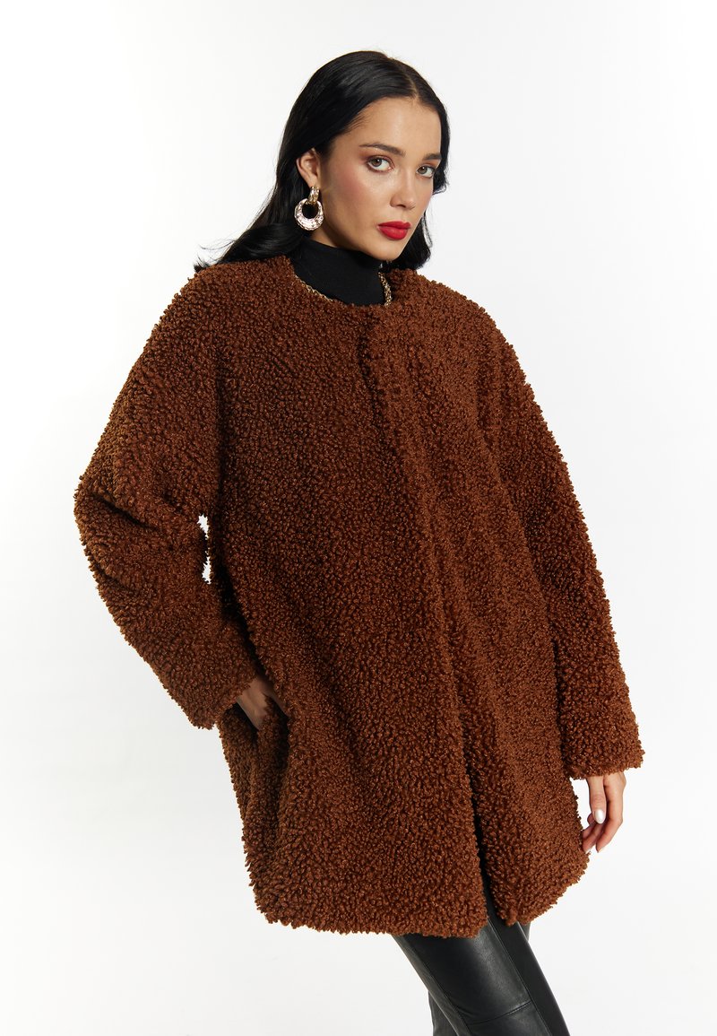 Brun, fluffy, tekstureret frakke med løs pasform, rund halsudskæring og sidelommer. Bæres over en sort turtleneck. Glat, skinnende stof i bunden.