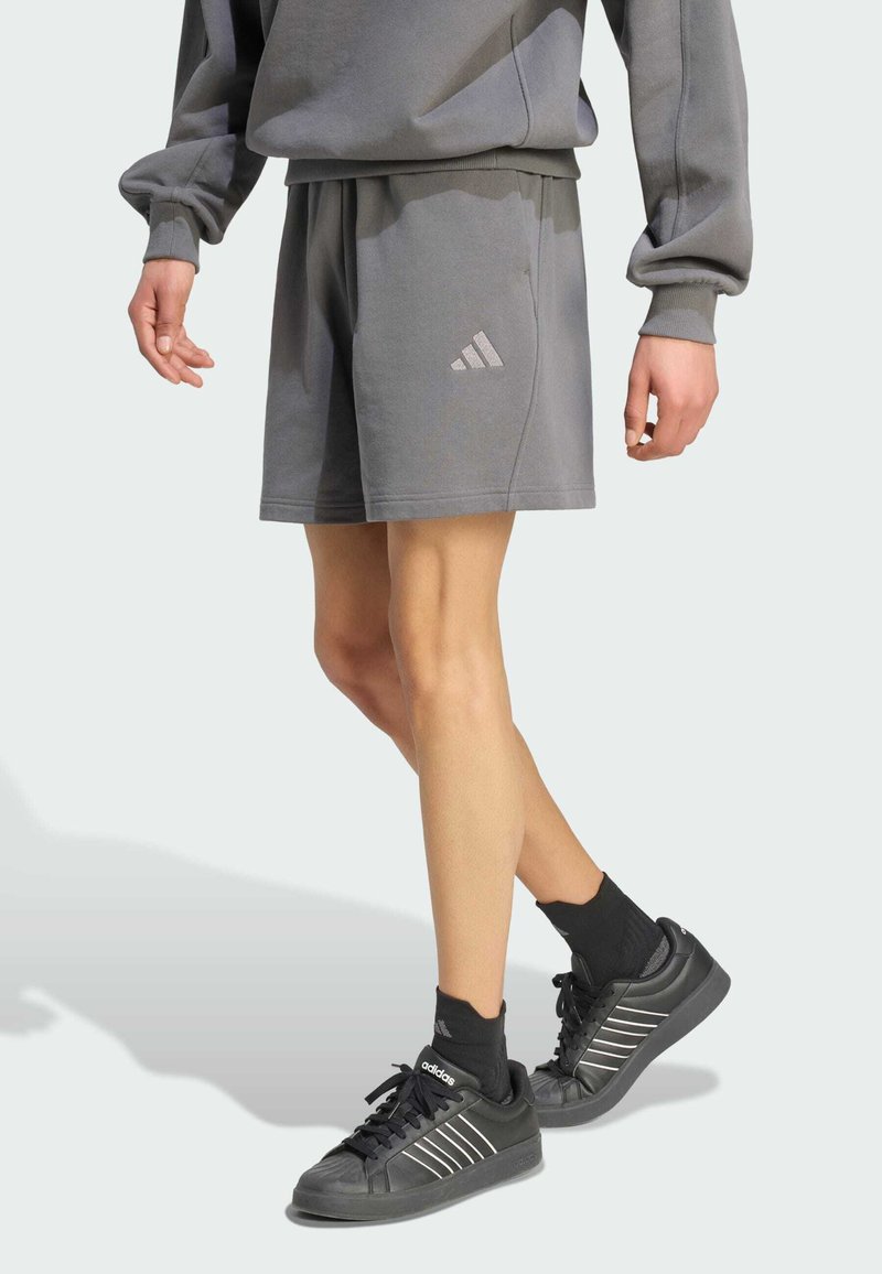 Personne portant un short gris Adidas, un haut à manches longues assorti, des chaussettes noires Adidas et des baskets noires Adidas, debout, jambes croisées.