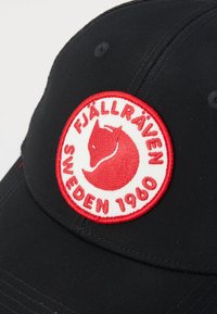 Svart caps med rund krone som har en fremtredende rød og hvit sirkulær lapp med et stilisert rev-logo og tekst.
