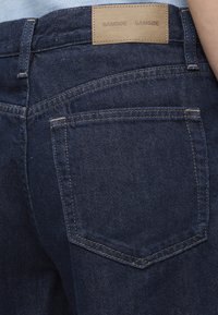Poche arrière d'un jean en denim bleu foncé avec une étiquette de marque en cuir beige "SAMSOE SAMSOE" cousue au-dessus de la poche.