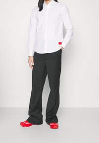 Vit långärmad skjorta med knapp, med en röd etikett, i kombination med löst sittande svarta byxor och klara röda skor med en texturerad design.