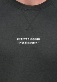 Musta collegepaita, jossa pyöreä pääntie ja raglan-hihat, valkoisella kirjailulla teksti "CRAFTED GOODS - PIER ONE DENIM." Pehmeä pinta.