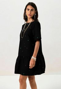 Schwarzes Kurzkleid mit rüschenbesäumtem Saum, bestickt mit bunten Blumenmustern. Der Stoff wirkt weich und hat eine lockere Passform.