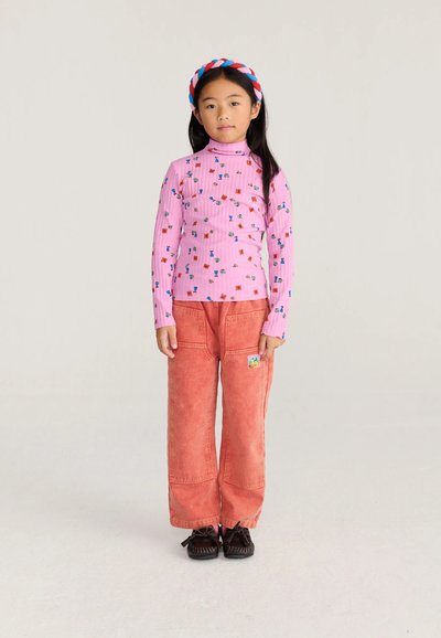 Fille portant un col roulé fleur rose, un pantalon corail à jambes larges, des chaussures noires et un bandeau tressé rouge, blanc et bleu, debout sur un fond uni.