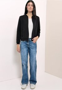 Zwarte bomberjack met rits en geribbelde textuur, gecombineerd met lichtblauwe wijde jeans en witte sneakers. Model staat met handen in de zij.