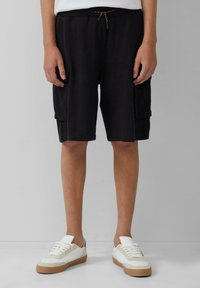 Schwarze Shorts mit Seitentaschen, elastischem Bund und oranger Kordel. Getragen mit weißen Sneakers mit braunen Sohlen. Glatte Stoffoberfläche.