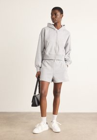 Felpa grigia con zip frontale e cappuccio, abbinata a pantaloni corti coordinati. Sneakers bianche e una piccola borsa nera completano l'outfit.