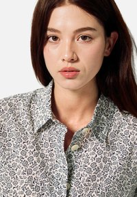 Mladá žena s hnědými vlasy a hnědýma očima, oblečená v bílé košili s černým květinovým vzorem, hledící přímo před sebe.