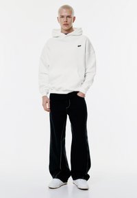 Vit huva tröja med framficka och svart logga, kombinerad med lösa svarta byxor och vita sneakers. En enkel, avslappnad design.