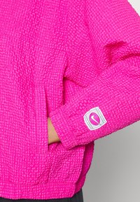 Veste légère de couleur rose vif avec une surface texturée, présentant un poignet élastique et un petit patch logo sur la manche.