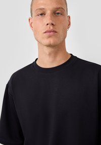 Jauns vīrietis ar īsiem matiem, tērpies vienkāršā melnā T-kreklā ar apaļu kakla izgriezumu, uz kura krūtīm ir mazs izšūts "QS" logo, uz vienkārša fona.