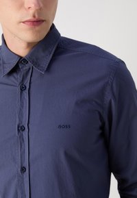 Cămașă bleu marin, cu nasturi, confecționată dintr-un material neted. Prezintă un guler clasic, nasturi în ton cu țesătura și logo-ul brodat "BOSS" pe pieptul stâng.