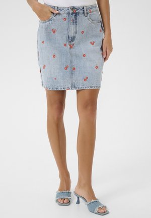 Jupe en jean bleu clair avec imprimé cerises rouges portée avec des sandales à talons effilochées bleu clair sur une personne debout.