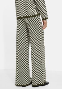 Pantalons en tricot à carreaux verts et blancs avec une coupe ample, présentant des jambes larges et une texture lisse. Des chaussures noires unies complètent la tenue.