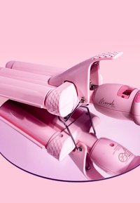Fer à friser rose avec une prise texturée, des accents en forme de diamant et un corps arrondi. Réfléchi sur une surface miroir circulaire.