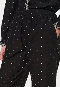 Svart tyg med ett vitt prickigt mönster, elastisk midja och dekorativt kantband vid ärmsluten. Mjuk textur, avspänd passform.
