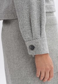 Main droite posée sur le côté, portant une chemise à manches longues à rayures grises avec des poignets boutonnés et une jupe ou un pantalon assorti.