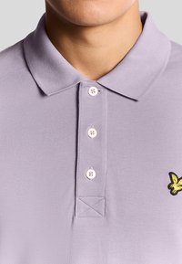 Polo shirt di colore viola chiaro con una trama testurizzata, tre bottoni e un logo di uccello ricamato giallo sul lato sinistro.