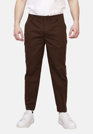 Uomo che indossa pantaloni slim marrone scuro e sneakers bianche, mostrato dalla vita ai piedi su uno sfondo semplice.