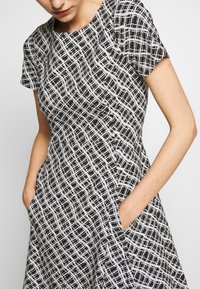 Femme portant une robe à carreaux noir et blanc à manches courtes, les mains dans les poches, regardant légèrement vers le bas.