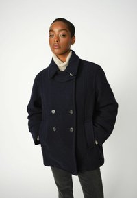 Cappotto doppiopetto di lana blu navy con colletto alto, due tasche frontali e dettagli con bottoni, abbinato a un maglione di colore chiaro sotto.