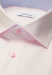 Col de chemise texturée rose clair avec devant assorti et boutons assortis, avec une étiquette "seidensticker" visible à l'intérieur du col.