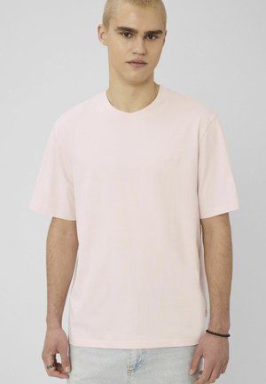 Jeune homme aux cheveux blonds courts portant un t-shirt rose clair et un jean bleu clair, debout devant un fond gris uni.