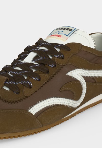 Καφέ αθλητικό παπούτσι με σκούρα καφέ κορδόνια, λευκό ραφή ζιγκ-ζαγκ και λευκή ετικέτα στη γλώσσα με την ένδειξη "Sandro Light Wave."
