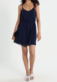 Vestido sin mangas color azul marino con tirantes ajustables, cintura elástica y falda fluida. Combinado con sandalias plateadas con tiras y un bolso negro de mano.